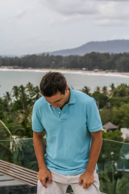 2011.05.10, Phuket, Tayland. Otelin çatısında cam çitlerin yanında duran genç bir adam. Phuket plajının arka planında kameraya bakan bir adam.