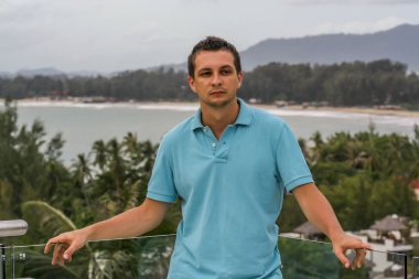 2011.05.10, Phuket, Tayland. Otelin çatısında cam çitlerin yanında duran genç bir adam. Phuket plajının arka planında kameraya bakan bir adam.