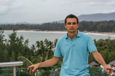 2011.05.10, Phuket, Tayland. Otelin çatısında cam çitlerin yanında duran genç bir adam. Phuket plajının arka planında kameraya bakan bir adam.