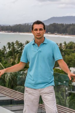 2011.05.10, Phuket, Tayland. Otelin çatısında cam çitlerin yanında duran genç bir adam. Phuket plajının arka planında kameraya bakan bir adam.