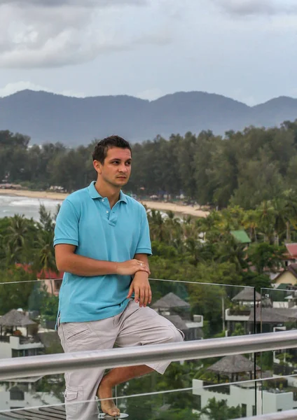 2011.05.10, Phuket, Tayland. Otelin çatısında cam çitlerin yanında duran genç bir adam. Phuket plajının arka planında kameraya bakan bir adam.