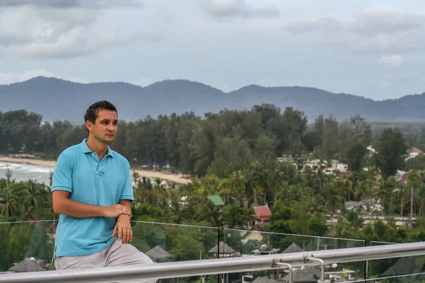 2011.05.10, Phuket, Tayland. Otelin çatısında cam çitlerin yanında duran genç bir adam. Phuket plajının arka planında kameraya bakan bir adam.