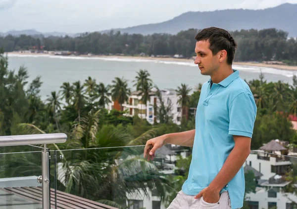 2011.05.10, Phuket, Tayland. Otelin çatısında cam çitlerin yanında duran genç bir adam. Phuket plajının arka planında kameraya bakan bir adam.