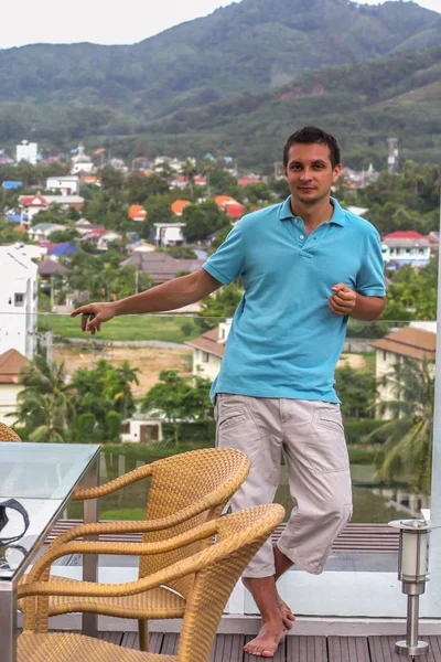 2011.05.10, Phuket, Tayland. Otelin çatısında cam çitlerin yanında duran genç bir adam. Phuket plajının arka planında kameraya bakan bir adam.