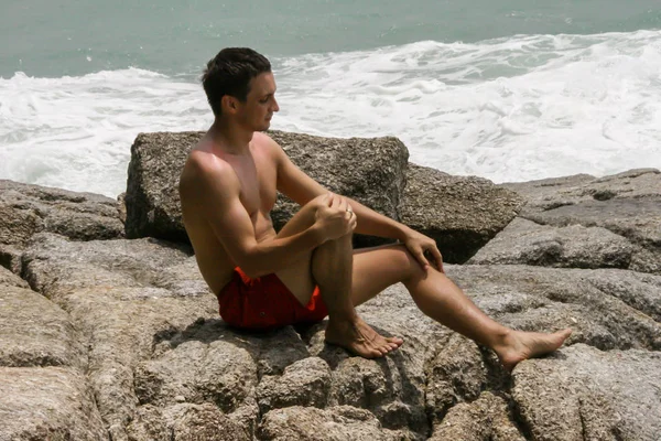 2011.05.10, Phuket, Tayland. Kırmızı yüzme şortu giyen genç bir adam kayaların üzerinde oturmuş okyanusa bakıyor. Yaz sahilde istirahat. 