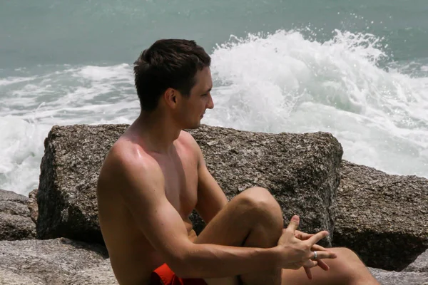 2011.05.10, Phuket, Tayland. Kırmızı yüzme şortu giyen genç bir adam kayaların üzerinde oturmuş okyanusa bakıyor. Yaz sahilde istirahat. 