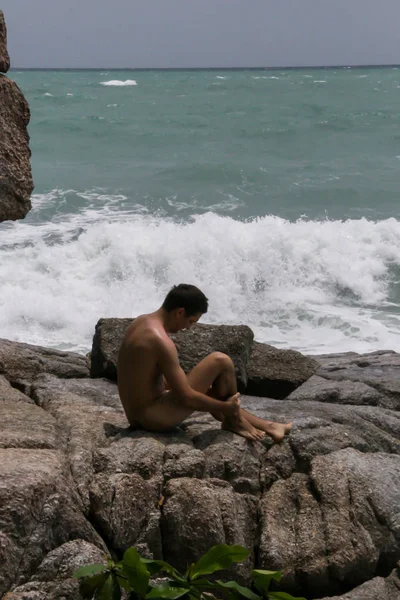 2011.05.10, Phuket, Tayland. Genç bir çıplak adam kayaların üzerinde oturuyor ve kameraya bakıyor. Yaz sahilde istirahat. 