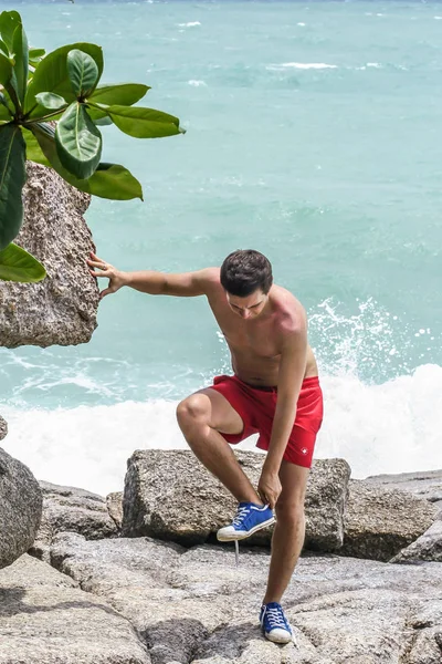2011.05.10, Phuket, Tayland. Kırmızı yüzme şortu giyen genç bir adam spor ayakkabılarını çıkarıyor. Kayalıklarda bir adam..