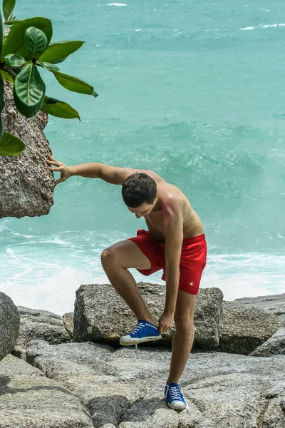 2011.05.10, Phuket, Tayland. Kırmızı yüzme şortu giyen genç bir adam spor ayakkabılarını çıkarıyor. Kayalıklarda bir adam..