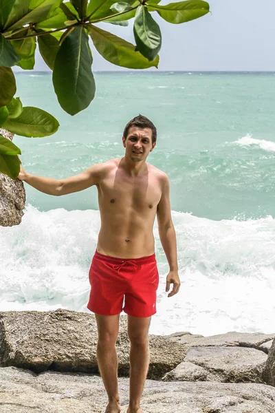 2011.05.10, Phuket, Tayland. Kırmızı yüzme şortu giyen genç bir adam kayaların üzerinde durup okyanusa bakıyor. Yaz sahilde istirahat. 