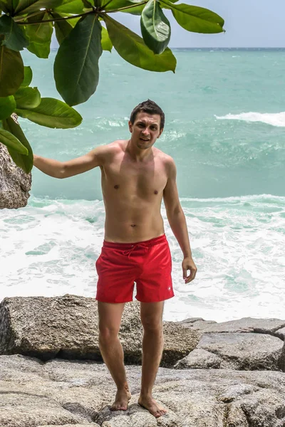 2011.05.10, Phuket, Tayland. Kırmızı yüzme şortu giyen genç bir adam kayaların üzerinde durup okyanusa bakıyor. Yaz sahilde istirahat. 