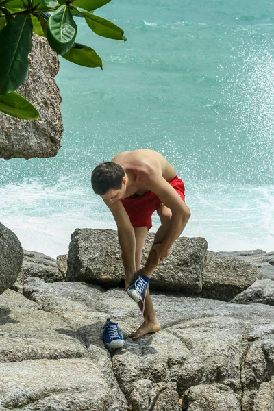 2011.05.10, Phuket, Tayland. Kırmızı yüzme şortu giyen genç bir adam spor ayakkabılarını çıkarıyor. Kayalıklarda bir adam..