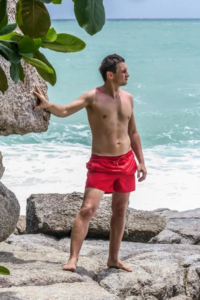 2011.05.10, Phuket, Tayland. Kırmızı yüzme şortu giyen genç bir adam kayaların üzerinde durup okyanusa bakıyor. Yaz sahilde istirahat. 
