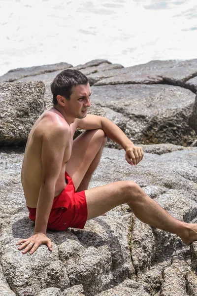 2011.05.10, Phuket, Tayland. Kırmızı yüzme şortu giyen genç bir adam kayaların üzerinde oturmuş okyanusa bakıyor. Yaz sahilde istirahat. 