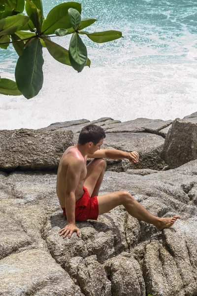 2011.05.10, Phuket, Tayland. Kırmızı yüzme şortu giyen genç bir adam kayaların üzerinde oturmuş okyanusa bakıyor. Yaz sahilde istirahat. 