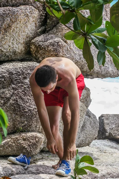 2011.05.10, Phuket, Tayland. Kırmızı yüzme şortu giyen genç bir adam spor ayakkabılarını çıkarıyor. Kayalıklarda bir adam..