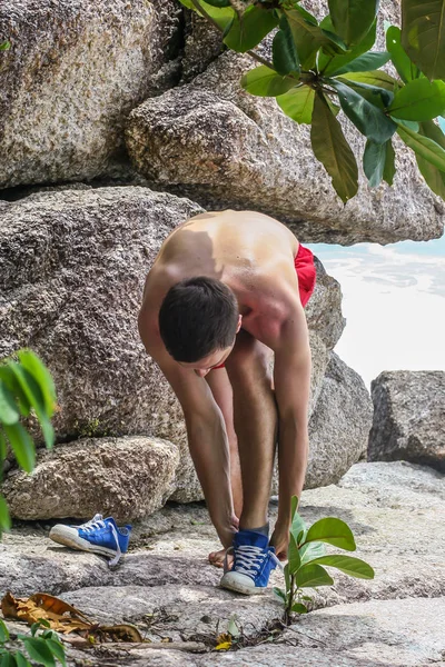2011.05.10, Phuket, Tayland. Kırmızı yüzme şortu giyen genç bir adam spor ayakkabılarını çıkarıyor. Kayalıklarda bir adam..