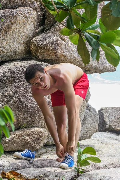 2011.05.10, Phuket, Tayland. Kırmızı yüzme şortu giyen genç bir adam spor ayakkabılarını çıkarıyor. Kayalıklarda bir adam..