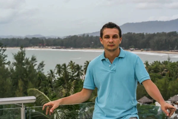 2011.05.10, Phuket, Tayland. Otelin çatısında cam çitlerin yanında duran genç bir adam. Phuket plajının arka planında kameraya bakan bir adam.