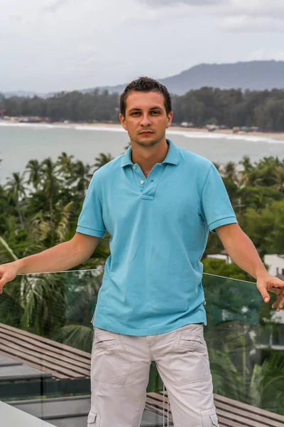 2011.05.10, Phuket, Tayland. Otelin çatısında cam çitlerin yanında duran genç bir adam. Phuket plajının arka planında kameraya bakan bir adam.