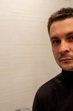 Işığın arka planında siyah balıkçı yaka giyen ve kameraya bakan kısa saçlı genç bir adamın yarım portresi..