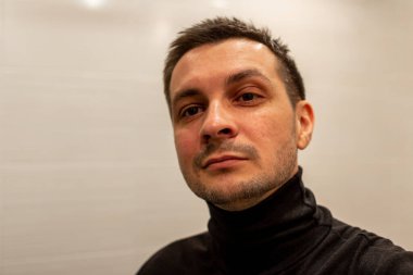 Arka planda siyah balıkçı yaka giyen ve kameraya bakan kısa saçlı genç bir adamın portresi..