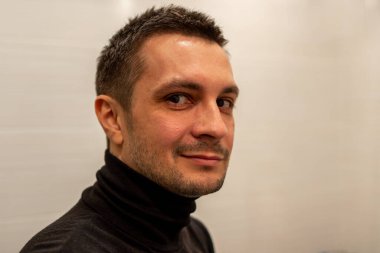 Arka planda siyah balıkçı yaka giyen ve kameraya bakan kısa saçlı genç bir adamın portresi..