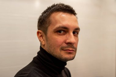 Arka planda siyah balıkçı yaka giyen ve kameraya bakan kısa saçlı genç bir adamın portresi..