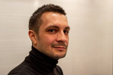 Arka planda siyah balıkçı yaka giyen ve kameraya bakan kısa saçlı genç bir adamın portresi..