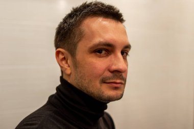 Arka planda siyah balıkçı yaka giyen ve kameraya bakan kısa saçlı genç bir adamın portresi..