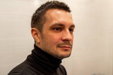 Siyah balıkçı yaka kazak giyen ve arka plandaki ışığa bakan kısa saçlı bir adamın portresi..