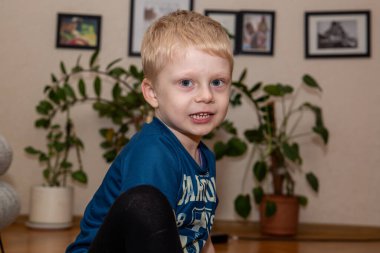 Küçük sarışın bir çocuk ev bitkilerinin arka planında oturuyor ve duvarda fotoğraf var (kameraya bakıyor)).