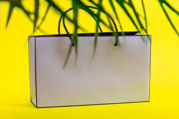 White paper bag and palm tree leaf on yellow background. Concept of shopping.