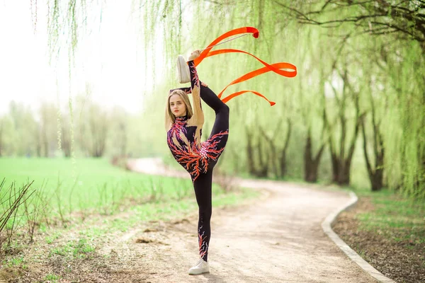 Rhythmic gymnastics. Young gymnast girl with red ribbon outdoor in park