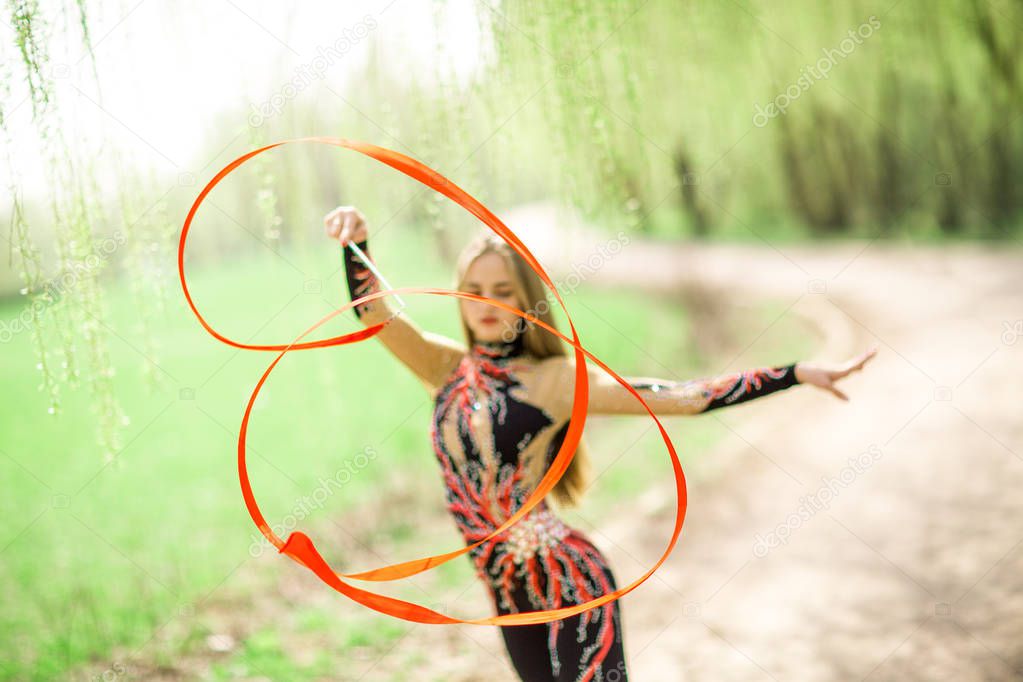 Rhythmic gymnastics. Young gymnast girl with red ribbon outdoor in park