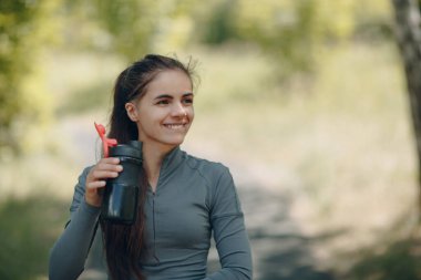 Smiling runner woman jogger drinking bottled water after jogging in park outdoor.