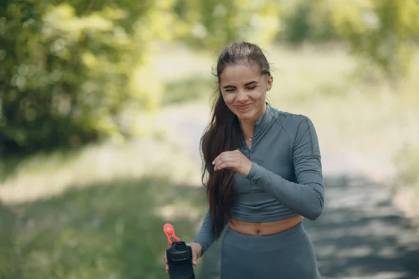 Smiling woman jogger in gray tracksuit drinking bottled water after jogging in park outdoor. — Stok fotoğraf
