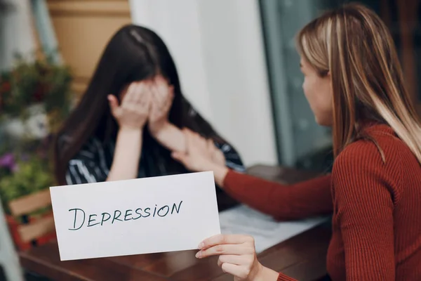 Verborgen depressie concept. Een huilende vrouw. Tweede vrouw met wit vel papier met het label Depressie in de hand. Twee vrouwen aan het praten in een café. — Stockfoto