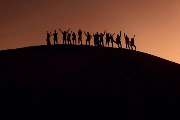 Persone in piedi sulla cima di dune nel deserto con le mani in alto. Silhouette al tramonto. — Foto Stock