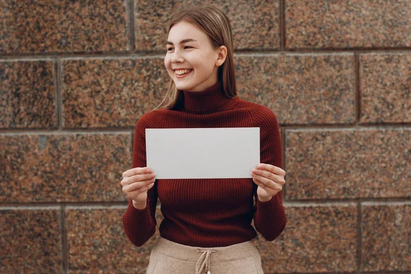 Mujer joven sonriendo con sonrisa sostiene el papel blanco en el fondo de piedra de la mano. Chica con hoja de plantilla en blanco con espacio vacío . — Foto de Stock