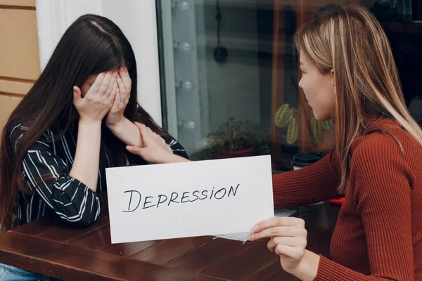 Versteckte Depression. Frau weint. Eine zweite Frau hält ein weißes Blatt Papier mit der Aufschrift Depression in der Hand. Zwei Frauen unterhalten sich in Straßencafé. — Stockfoto