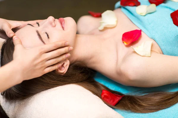 Young beautiful woman receiving head face massage in beauty spa with rose flower petals. — Stock Photo, Image
