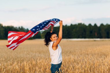 Gün batımında buğday tarlasında Amerikan bayrağı taşıyan bir kadın. 4 Temmuz. Bağımsızlık Günü Vatanseverlik Günü.