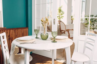 Mutfak masasında yeşil bardaklar ve beyaz tabaklar. Arka planda yeşil bitki var..