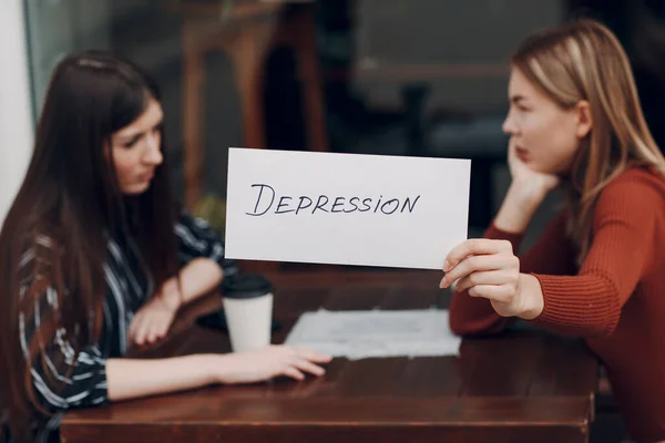 Frau mit weißem Blatt Papier mit der Aufschrift Depression in der Hand. Zwei Frauen unterhalten sich in einem Straßencafé. Verstecktes Depressionskonzept — Stockfoto