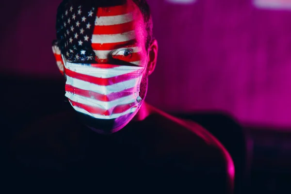 Pria dewasa bertopeng wajah medis dengan bendera Amerika Serikat di wajahnya dalam gelap. — Stok Foto