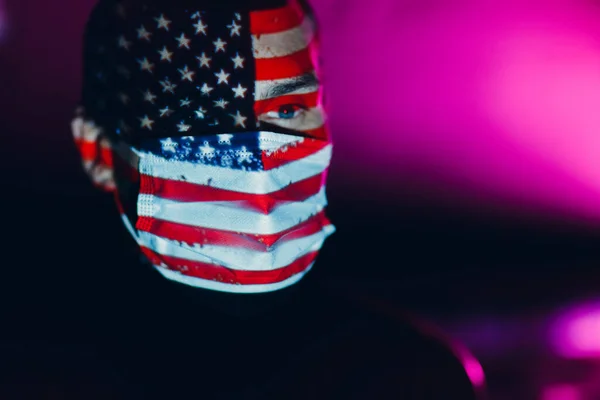 Hombre adulto en mascarilla médica con bandera de EE.UU. en su cara en la oscuridad. — Foto de Stock