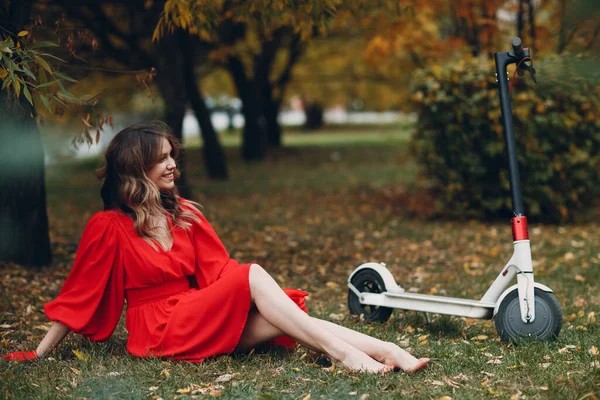Jovem mulher sentada com scooter elétrico em vestido vermelho no parque da cidade de outono — Fotografia de Stock
