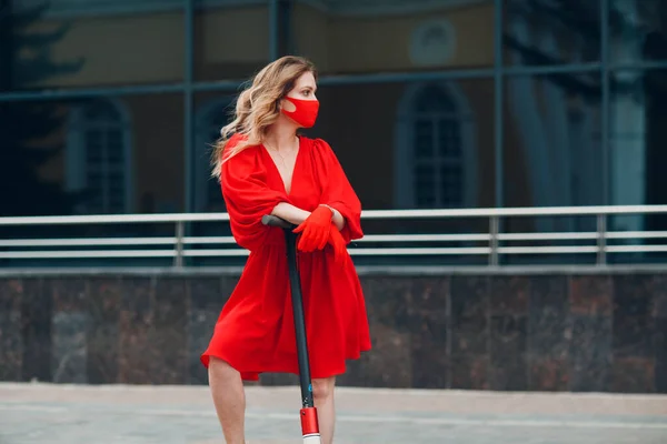 Junge Frau mit Elektroroller im roten Kleid und Handschuhen mit medizinischer Gesichtsmaske in der Stadt — Stockfoto