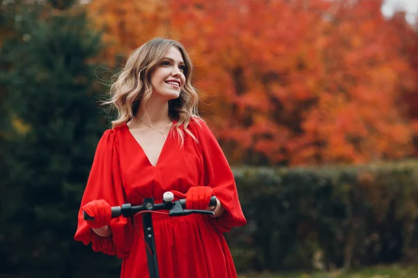 Молодая женщина, остановившаяся на электроскутере в красном платье и улыбающаяся в осеннем городском парке — стоковое фото